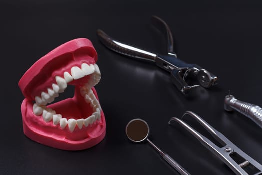Layout of a human jaw with the rubber dam forcep and the dental hole punch. Head of high-speed dental handpiece with bur, a metal mirror on the black background.