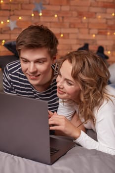 Smiling young man and woman lying in bed and watching something on the laptop. They're happy and lovely