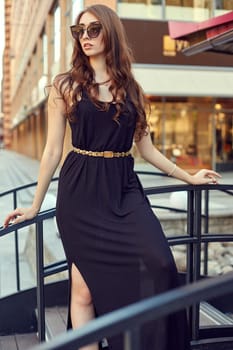 Fashion style portrait of young beautiful elegant woman in black dress, sunglasses, with long dark hair walking at city streets.