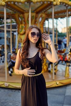 Portrait of a beautiful brunette woman in evening dress next to a children's carousel. Fashion style portrait of young beautiful elegant woman in black dress