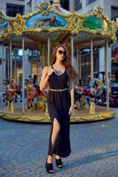 Portrait of a beautiful brunette woman in evening dress next to a children's carousel. Fashion style portrait of young beautiful elegant woman in black dress