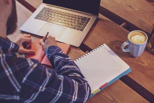 Cropped image of casual business man or freelancer planning his work on notebook, working on laptop computer with smart phone, cup of coffee on table at coffee shop or home office, working from cafe concept