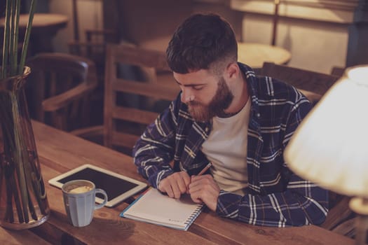 Young bearded businessman sits in cafe, home at table and writes in notebook, near lies tablet computer with black screen. Man is working, studying. Online education, marketing, training.
