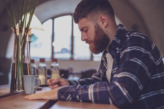Young bearded businessman sits in cafe, home at table and writes in notebook. Man is working, studying. Online education, marketing, training.