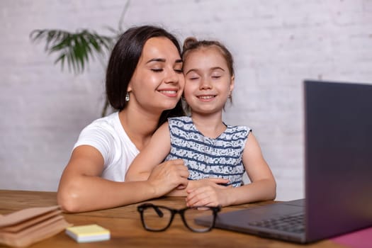 Attractive young woman and her little cute daughter are sitting at the table and having fun while doing homework together. Mother helps daughter with her school classes.