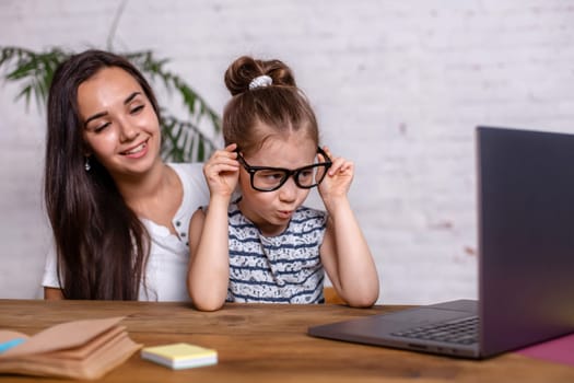 Attractive young woman and her little cute daughter are sitting at the table and having fun while doing homework together. Mother helps daughter with her school classes.