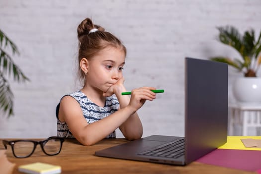 Cute little girl is sitting at table with her laptop and notebook, wearing glasses. The concept of new programs for teaching children