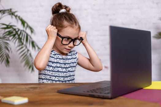 Cute little girl is sitting at table with her black laptop and wearing glasses at home. A child girl doing homework or playing game