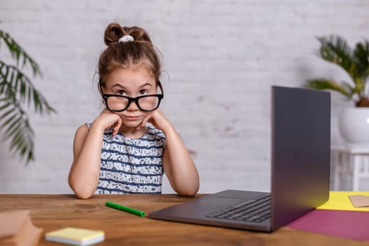 Cute little girl is sitting at table with her laptop and notebook, wearing glasses. The concept of new programs for teaching children