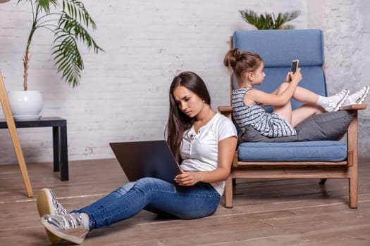 Pretty brunette mother working with laptop and cute little girl laying on the armchair and playing games on the smartphone at home
