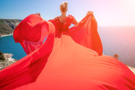 woman sea red dress. Blonde with long hair on a sunny seashore in a red flowing dress, back view, silk fabric waving in the wind. Against the backdrop of the blue sky and mountains on the seashore