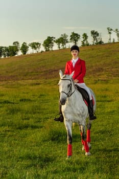 Young woman rider, wearing red redingote and white breeches, with her horse in evening sunset light. Outdoor photography in lifestyle mood