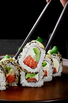 Delectable Philadelphia rolls in sesame stuffed with salmon, cream cheese, cucumber and avocado topped with mayonnaise and greens served on plate. One roll grasped by chopsticks. Popular food concept