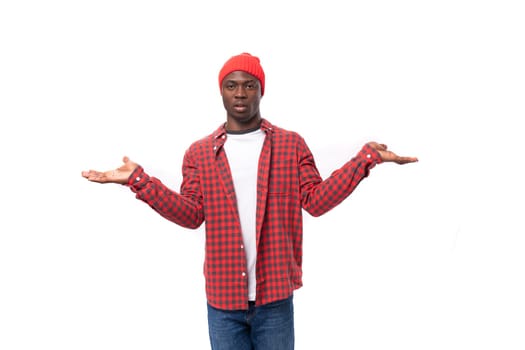 portrait of confused 30s black american man dressed in red shirt and cap shrugging against white studio background with copy space.