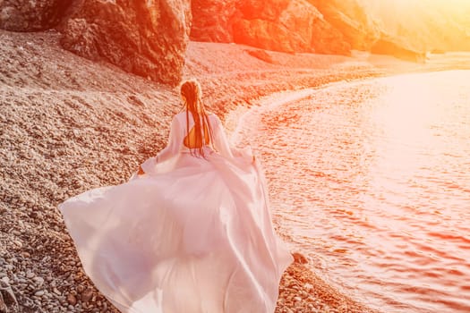 Mysterious woman silhouette long hair walks on the beach ocean water, sea nymph wind listens to the wave. Throws up a long white dress, a divine sunset. Artistic photo from the back without a face.