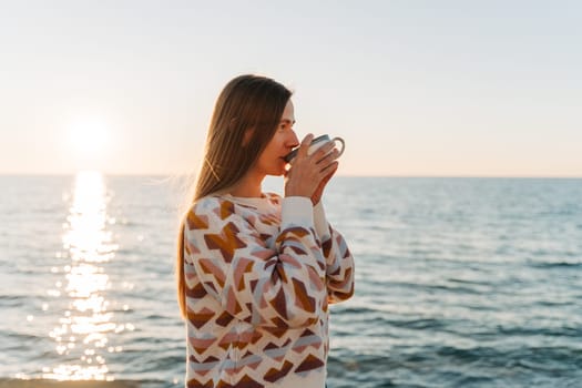 Young beautiful girl in cozy sweater drinking coffee while enjoying winter sun on seaside shore during mild sunset. Cute attractive woman enjoying cup of tea with autumn ocean sun trail background.