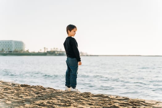 School boy child enjoying winter sea view from a sandy beach. Kid standing on shore watching autumn ocean waves