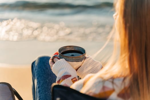 Closeup of a woman sitting in chair on winter sea shore in a cozy sweater and holding hot cup of coffee. A girl enjoying beautiful view of autumn ocean seaside waterfront waves.