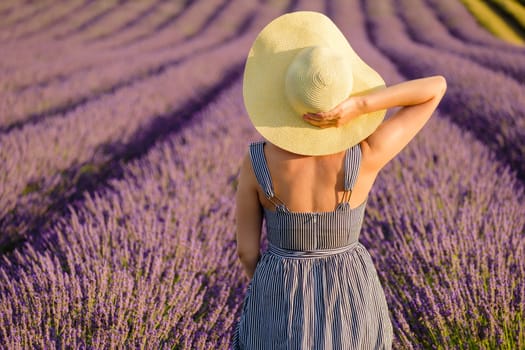 In the midst of the picturesque lavender field, a young lady donning a sundress gracefully rests her hands on her straw hat, savoring the breathtaking nature surrounding her on a sunlit summer day.