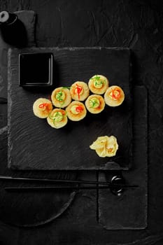 Top view of baked maki sushi rolls topped with melted cheese, japanese mayonnaise drops, colorful thin shaving of scallions and chili pepper served with gari and soy sauce on black slate board