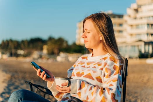 Young beautiful woman holding texting smartphone cellphone while drinking coffee on winter beach with sea waves in background. Cute attractive girl using mobile when enjoying tea on autumn seashore.