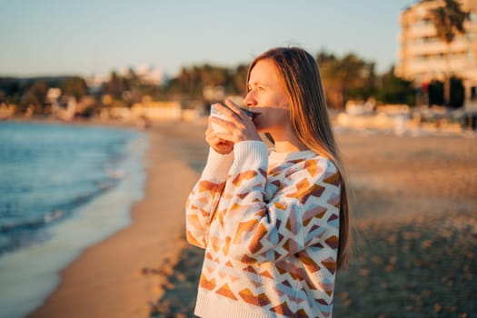 Young beautiful girl in cozy sweater drinking coffee on a winter seaside shore while taking sunbathe in a mild sunset. Cute attractive woman enjoying her cup of tea while watching autumn ocean waves.