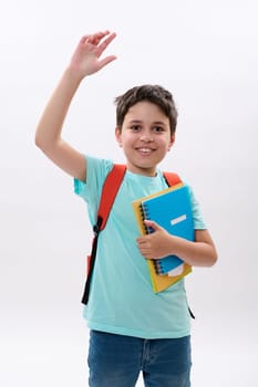 Emotional cheerful happy kid, smart schoolboy greeting waving hand, smiling looking at camera, carrying orange backpack and school supplies, isolated on white background. Education. Learning. Studying