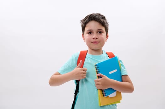 Caucasian smart school child, handsome preteen boy in blue t-shirt, carrying school supplies and orange backpack, smiles looking at camera, thumbs up, shows approval sign. Isolated white background