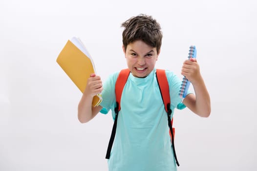 Caucasian emotional teenage boy, confused angry schoolkid holding textbooks, expressing anger, irritation and frustration, isolated on white studio background. Problems at school and education concept