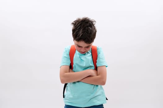 Offended distraught schoolboy with backpack, folding arms, standing tearful and looking down after bullying act at school, isolated on white background. Social issues. Problems. Puberty difficult age