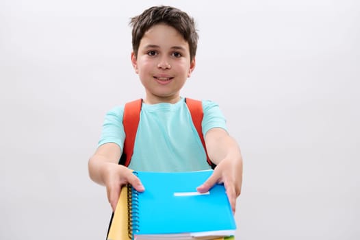 Smart smiling schoolboy carrying orange backpack, holding out at camera colorful textbooks and school supplies, isolated on white studio background. Copy ad space. Education. Learning. Back to school