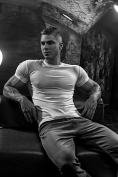 Young sexy man bodybuilder athlete, studio portrait in loft, guy model in white t-shirt and jeans against the background of stylized wall sits on leather sofa black and white photo