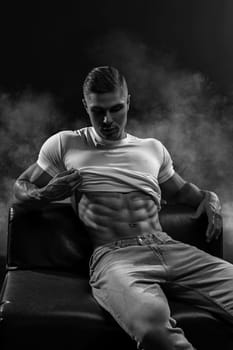 Young sexy man bodybuilder athlete, studio portrait in a loft, guy model shows abs sits on a leather sofa black and white photo
