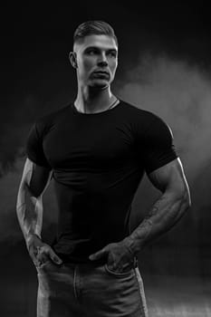 Muscular model sports young man in jeans and black t-shirt on black background. Fashion portrait of brutal sporty healthy strong muscle guy with a modern trendy hairstyle. Black and white photo