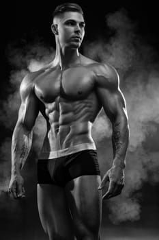 Bodybuilder adult muscular male model posing on black background in shorts demonstrates perfect abs with a low percentage of fat, black and white photo