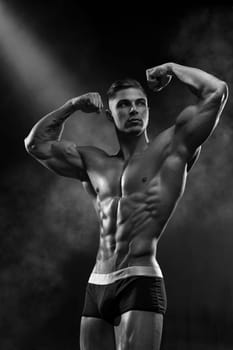 Muscular, chiselled man shows off a front double bicep, perfect six pack abs. Dramatic portrait of posing bodybuilder on black cyclorama, black and white photo