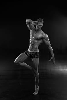 Athletic man in shorts dynamically poses while standing on one leg, purposefully leaning forward his body and covering his face with an open palm. Bodybuilder poses in an original way