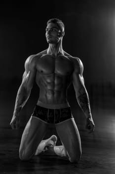 A strong athletic man poses shirtless standing on knees in black studio. Athletic muscular body with six pack abs and low percentage of fat black and white photo