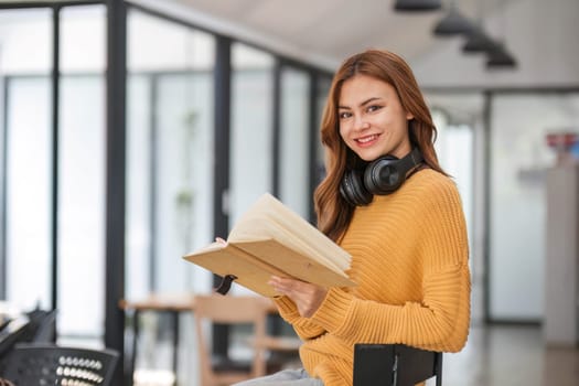 A portrait of an attractive young Asian female in a cozy yellow sweater stands inside a coffee shop with a book and headphones. college student, freelancer..