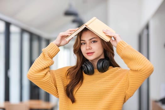 A portrait of an attractive young Asian female in a cozy yellow sweater stands inside a coffee shop with a book and headphones. college student, freelancer..