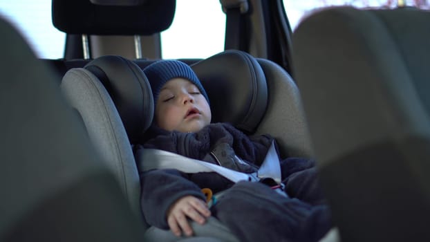 A small child sleeps in a car seat in a parking lot. Boy in a sweatshirt and hat. 4k