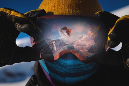 Close-up portrait of a skier in a mask holding on to ski goggles in the reflection of which the skier does a backflip trick. against the backdrop of snowy epic high mountains.