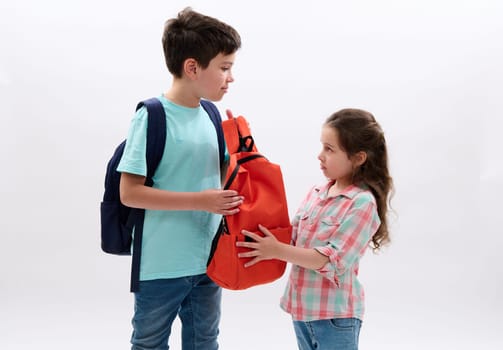 Caucasian preteen schoolboy with backpack carrying orange rucksack of his younger sister, ready to going for school, isolated over white background. Family relationships. Frienships. Help and support