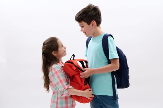 Caucasian happy children, preteen boy and preschooler little kid girl with backpacks, smile looking at each other, isolated over white studio background. Education. Back to school concept