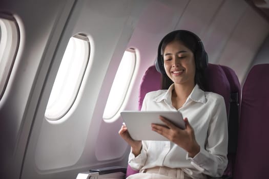 Successful beautiful young asian business woman sits in airplane cabin plane and works on digital tablet with stylus. Flying at first class.