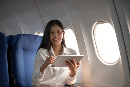 Successful beautiful young asian business woman sits in airplane cabin plane and works on digital tablet with stylus. Flying at first class.