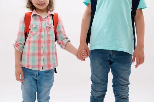 Closeup cute teen boy and preschooler girl, brother and younger sister holding hands, isolated on white. Family relationships. Happy childhood. Kids with backpack going to the school. Help. Support
