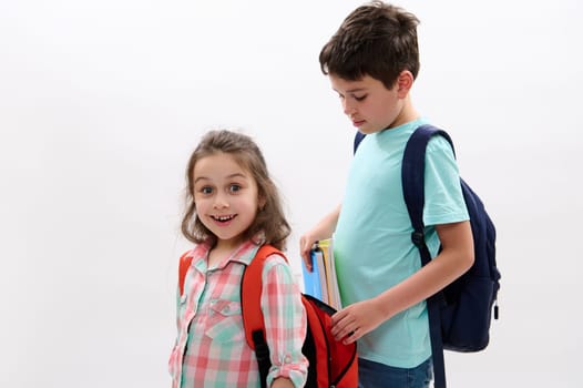 Lovely little kid girl, mischievous preschooler child smiles and makes faces looking at camera while her older brother puts workbooks and school supplies inside her backpack, over white background.