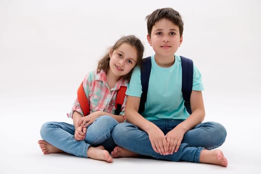 Authentic portrait of Caucasian teen boy and little girl, happy older brother and younger sister, school kids with backpacks, smiling at camera, sitting close to each other, isolated white background