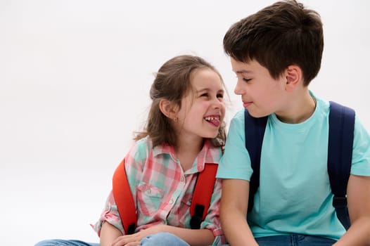 Mischievous little girl, shows tongue and makes faces, smiles to her older brother, isolated over white background. Cheerful smart school Kids with backpacks having fun together. Family relationships